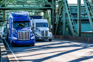 Blue big rig semi-truck with car hauler semi trailer and white big rig semi truck with dry van semi trailer driving side by side on the road on the truss draw bridge