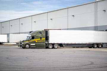 Green bonnet big rig semi truck with yellow strip with refrigerator semi trailer standing in the warehouse dock gate loading cargo for the next freight - obrazy, fototapety, plakaty