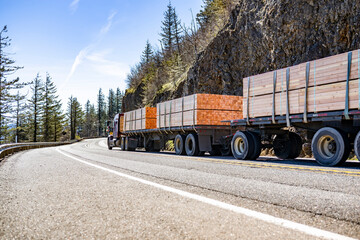 Powerful classic big rig semi truck tractor transporting fastened lumber wood on flat bed semi trailers running on the mountain road