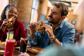 Young man enjoying in taste of hamburger while eating with friends in  pub.