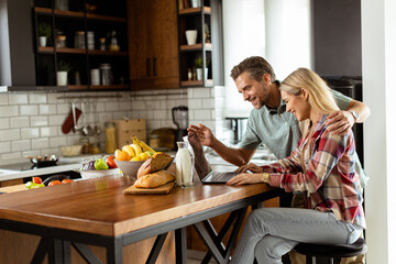 Cheerful couple enjoys a light-hearted moment in their sunny kitchen, working on laptop surrounded...