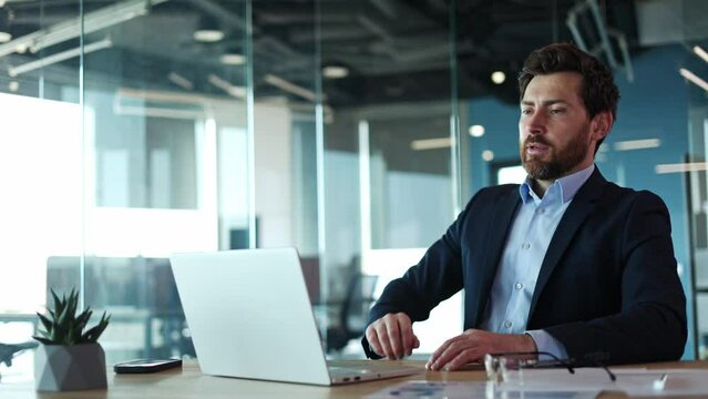 Exhausted man removing glasses and leaning back against chair backrest in office setting. Tired bearded caucasian businessman sitting in front wireless laptop and enjoying break for relax indoors.