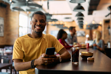 Happy black man using mobile phone while sitting in bar and looking at camera.