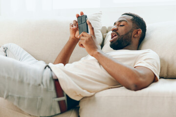 African American man sitting on a black sofa, happily using his mobile phone He is dressed in a white tshirt and is in a modern apartment The background shows a relaxed and comfortable lifestyle, with