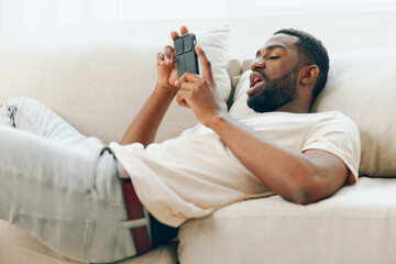 African American man sitting on a black sofa, happily using his mobile phone He is dressed in a...