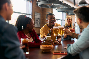 Cheerful black man toasting with his friends while drinking beer in pub. - obrazy, fototapety, plakaty