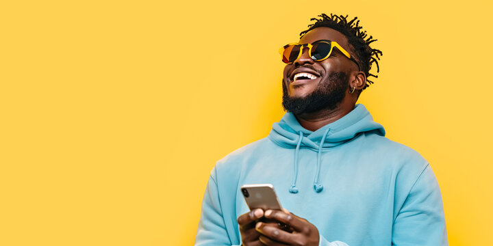 Happy African millennial man laughing with mobile phone on yellow studio backdrop. Joyful black man wearing blue hoodie, sunglasses smiling looking aside. Great new mobile App. People and gadgets