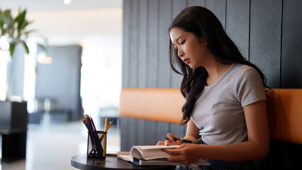 Asian teenage student woman reading a book and writing information from book to doing assignment homework for preparing test exam while studying and learning about education knowledge in cafe - 788990749