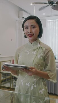 Vertical portrait of smiling female Asian spa salon manager working on digital tablet and looking at camera