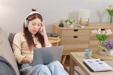 Asian business woman lying on sofa and wearing headphone to listening music during analyzing to planning about investment of new startup project and typing report on laptop while working from home - 788991147