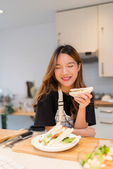 Young asian woman enjoying to having breakfast with eating delicious sandwich while cooking fresh vegetables salad and homemade snack for health food in modern kitchen with healthy lifestyle in home - 788991154