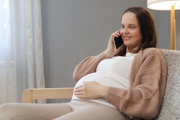 Beautiful pregnant woman sitting on sofa and using phone while calling to friend from home expressing positive emotions speaking about pregnancy and childbirth