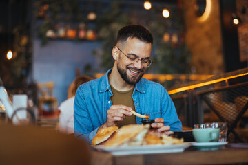 This is so delicious. Young man sitting in a cafe and enjoying in breakfast. Food, lifestyle concept. Happy young man having hamburger and french fries in fast food restaurant. - 788997140