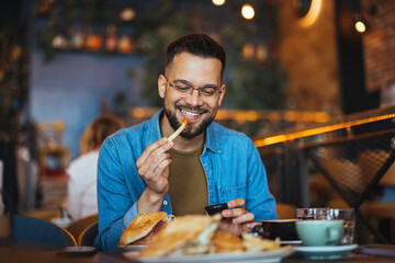 Portrait of a happy man eating at a restaurant and smiling - lifestyle concept. A young man with a beard sitting in a restaurant and holding hands french fries and going to eat - 788997372