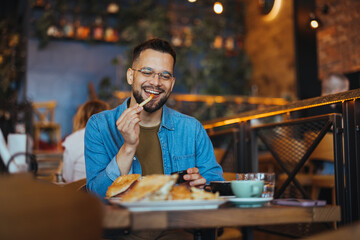 This is so delicious. Young man sitting in a cafe and enjoying in breakfast. Food, lifestyle concept. Happy young man having hamburger and french fries in fast food restaurant. - 788997532