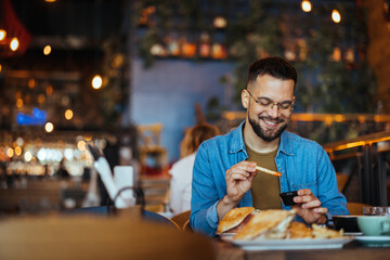Portrait of a happy man eating at a restaurant and smiling - lifestyle concept. A young man with a beard sitting in a restaurant and holding hands french fries and going to eat - obrazy, fototapety, plakaty