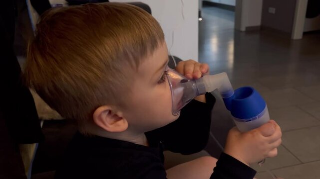 Close up of little caucasian sick boy holding a inhalation mask on his face to breath saline solution, at home