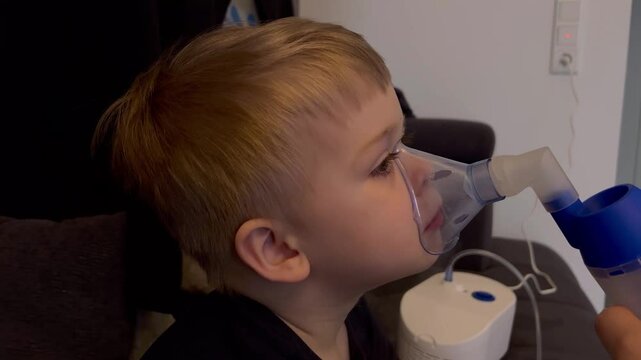 Close up of little caucasian sick boy holding a inhalation mask on his face to breath saline solution, at home