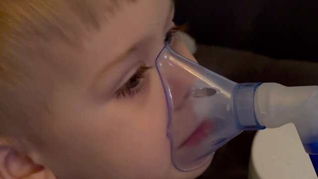 Close up of little caucasian sick boy holding a inhalation mask on his face to breath saline solution, at home