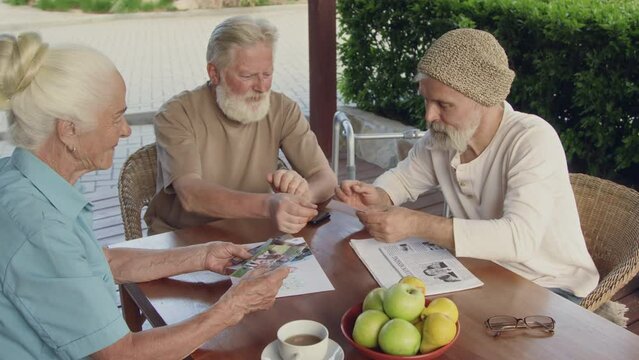 Medium shot of three assisted living facility residents sitting outdoors at table, elderly woman showing photos of grandchildren to male companions, man putting on glasses to look at picture