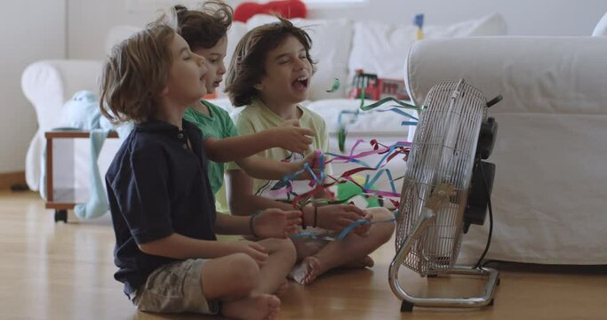 Three young boys playing with a spinning electric fan indoors during daytime