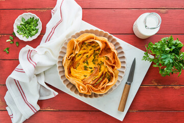 Homemade quiche or tart with slices of bacon and leeks with tortilla instead of dough on wooden cutting board on old red wooden background. Quiche open tart traditional French cuisine. Top view.