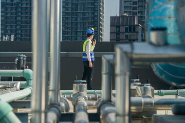 Focused engineer in hard hat and reflective vest standing amidst rooftop mechanical structures,...