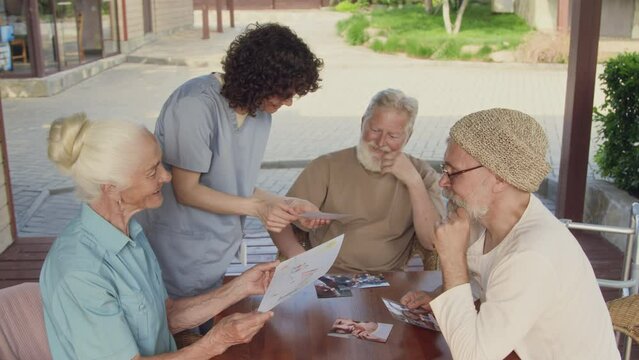 Medium shot of three elderly men and women and nurse sitting outdoors at table, admiring photographs of grandchildren and pencil drawing, chatting, sharing memories, while relaxing at retirement home