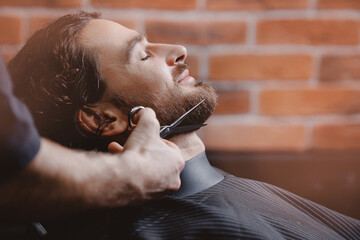Hipster man sitting in armchair barber shop while hairdresser shaves beard with scissors