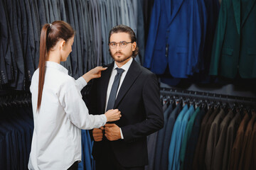 Beautiful woman seller smiling and help with choice suit to modern young handsome businessman. Shop...
