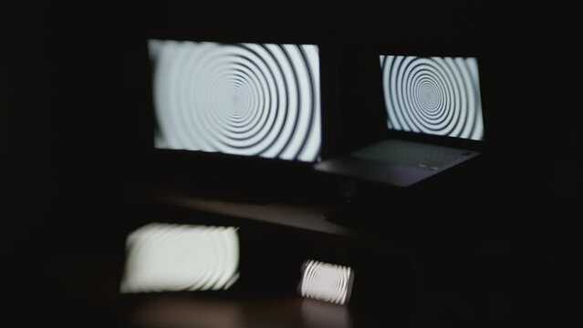 Angled shot of a hypnotic spiral pattern displayed on four device screens in a dark room with selective focus and a choppy shutter speed effect.
