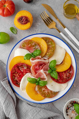 Salad of sliced multi-colored tomatoes, fresh peaches, mozzarella, prosciutto and basil on a gray...