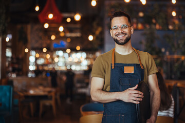 Portrait of Owner / Waiter at Restaurant. Portrait of handsome young male coffee shop owner standing at cafe. Portrait of a handsome barista in t-shirt and apron - 789034324