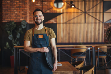 Waist up portrait of handsome waiter smiling cheerfully at camera standing in restaurant or cafe, copy space. Young waiter serving coffee in a cafe and looking at camera. - obrazy, fototapety, plakaty