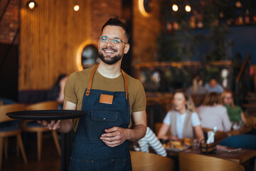 Happy waiter holding a tray while working in cafe and looking at camera. Young waiter serving coffee in a cafe and looking at camera. - 789035155