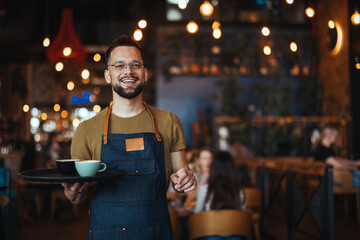 Portrait of smiling young waiter holding tray in cafe. Handsome waiter smiling at camera holding tray at the coffee shop. Profesional Waiter in Restaurant - 789036332