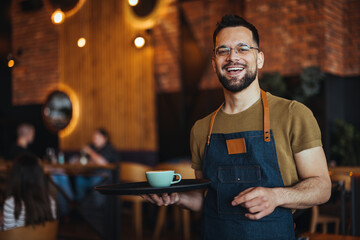 Portrait of smiling young waiter holding tray in cafe. Handsome waiter smiling at camera holding tray at the coffee shop. Profesional Waiter in Restaurant - 789036936