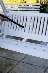 Man's hand cleaning a white garden rocking chair with a jet of pressurized water in the garden of...