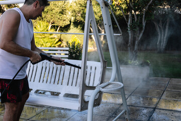 Casually dressed man in a bathing suit cleaning a seat in his backyard with a pressure washer on a summer afternoon. Summer concept and housework