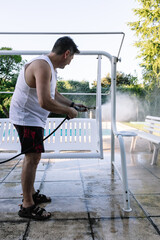 Fifty-year-old man outside his house, by the pool, cleaning a garden seat with a pressure washer, a...