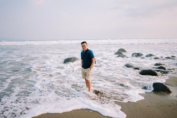 A contented man in a blue tee and shorts stands by the shore, his gaze fixed towards the horizon, radiating happiness against the backdrop of the beach