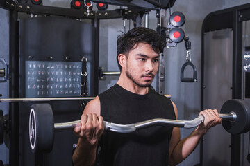 A fit asian man in a black tank top does basic standing barbell curls at the gym. Using an EZ curl bar with fixed weight to build biceps.