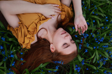 Young red haired woman in vintage dress laying on grass with blue flowers. Romantic top view...