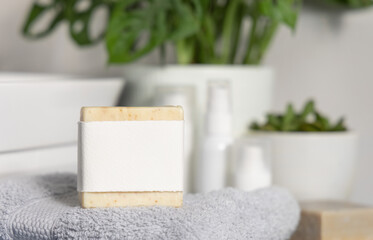 Beige Soap bar with blank label on light grey towel against green plant in bathroom, mockup