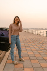 Vertical photo of asian Thai woman smiling and standing next to the car near the river at sunset time, chilling traveling on vocation. 