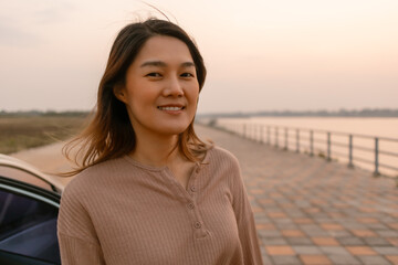 Photo of asian woman smiling and standing next to the car near the river at sunset time, chilling...