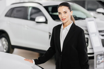 Portrait of a female car sales agent standing in front of new cars for sale in a modern car dealership. Automotive business concept.
