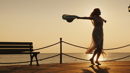A woman meets the dawn at sea. Emotionally spinning with a hat in his hand on the pier. Dream of travel concept
