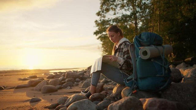Tourist woman looking a way on sea beach. Female having break in hike finding route look at paper map admiring nature at sunset sitting on stones. Camping female tourism travel wanderlust concept.