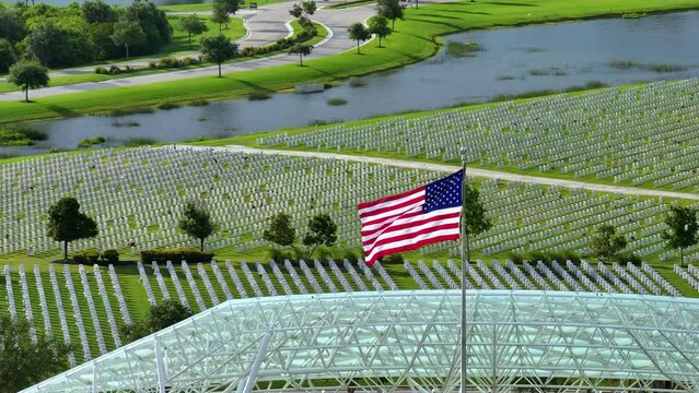 US national flag waving over military cemetery with rows of white tomb stones on green grass lawn. American Memorial Day concept